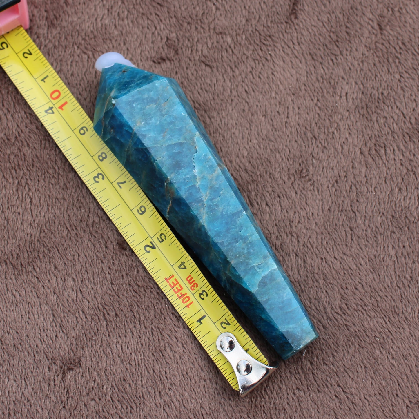 Blue Apatite Wand crystal measuring about 6 inches placed next to yellow tape measure on brown surface