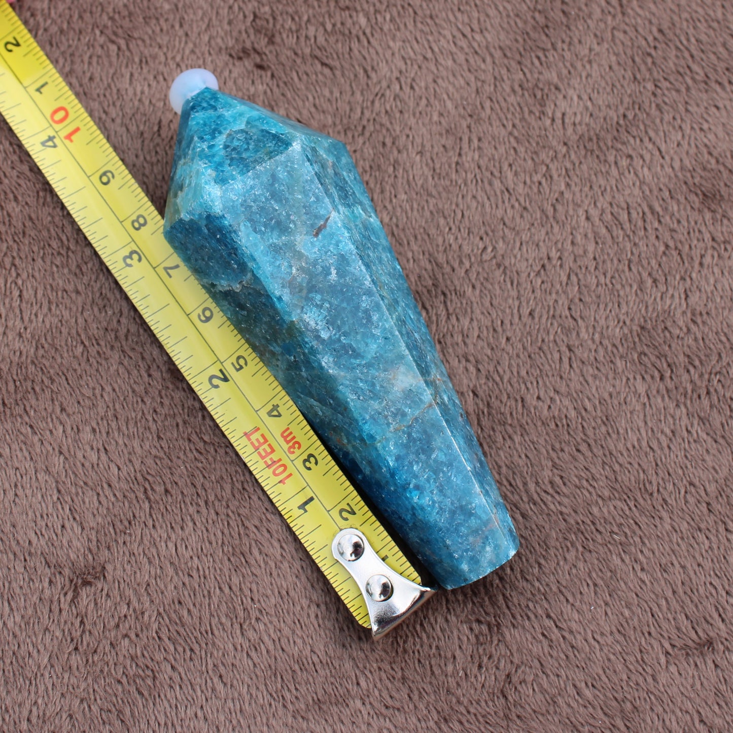Blue Apatite Wand crystal from Different Branches LLC shown next to yellow measuring tape on brown fabric