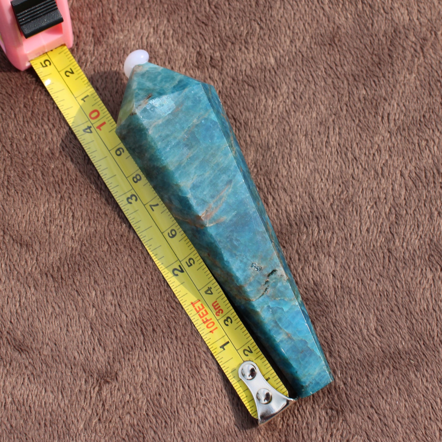 Blue Apatite Wand crystal placed next to a yellow measuring tape on a brown surface