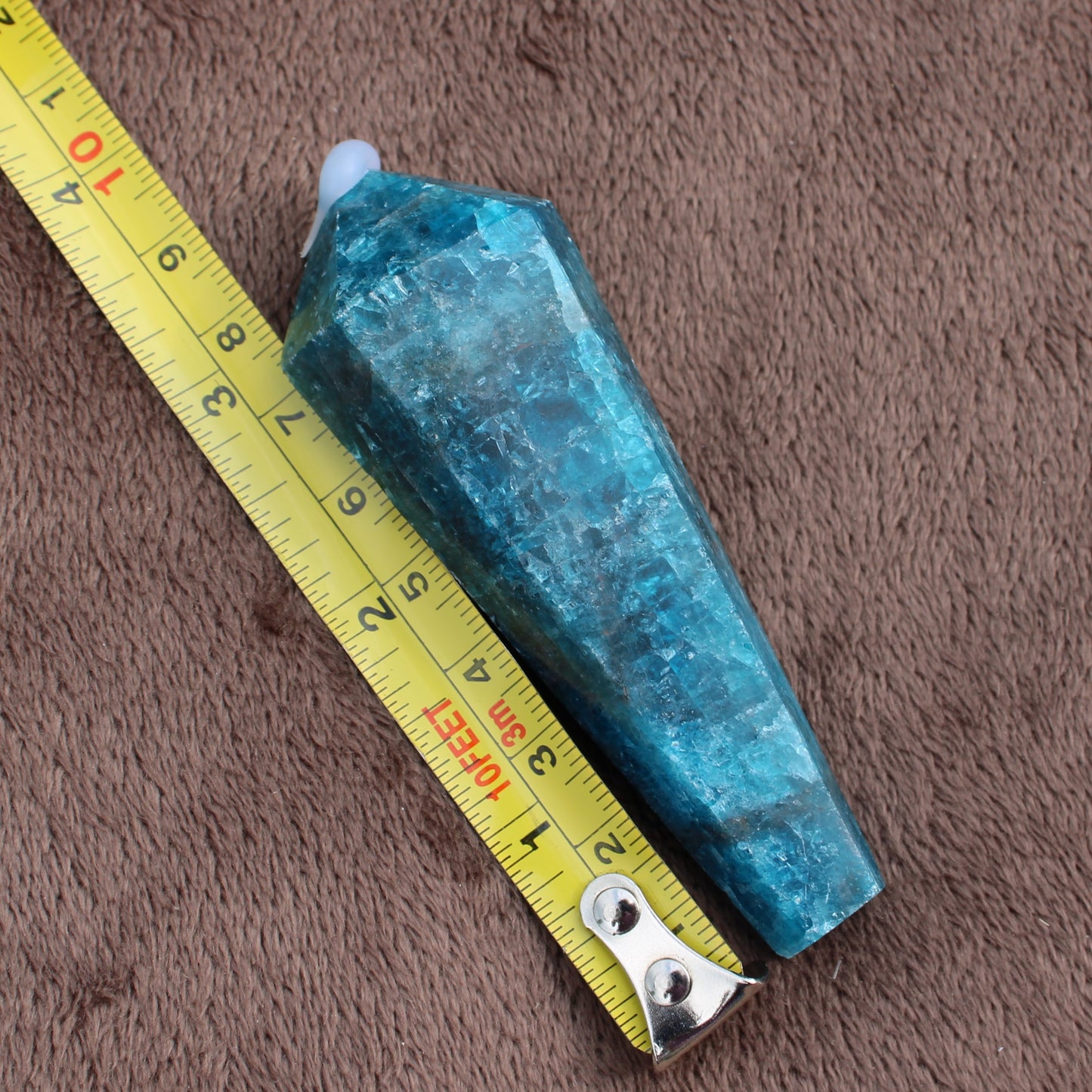 Blue Apatite Wand crystal measuring approximately 3 inches against a yellow tape measure on a brown textured surface