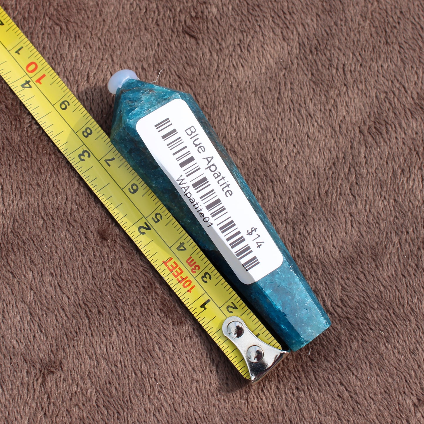 Blue Apatite Wand crystal from Different Branches LLC next to a measuring tape on a brown fabric background