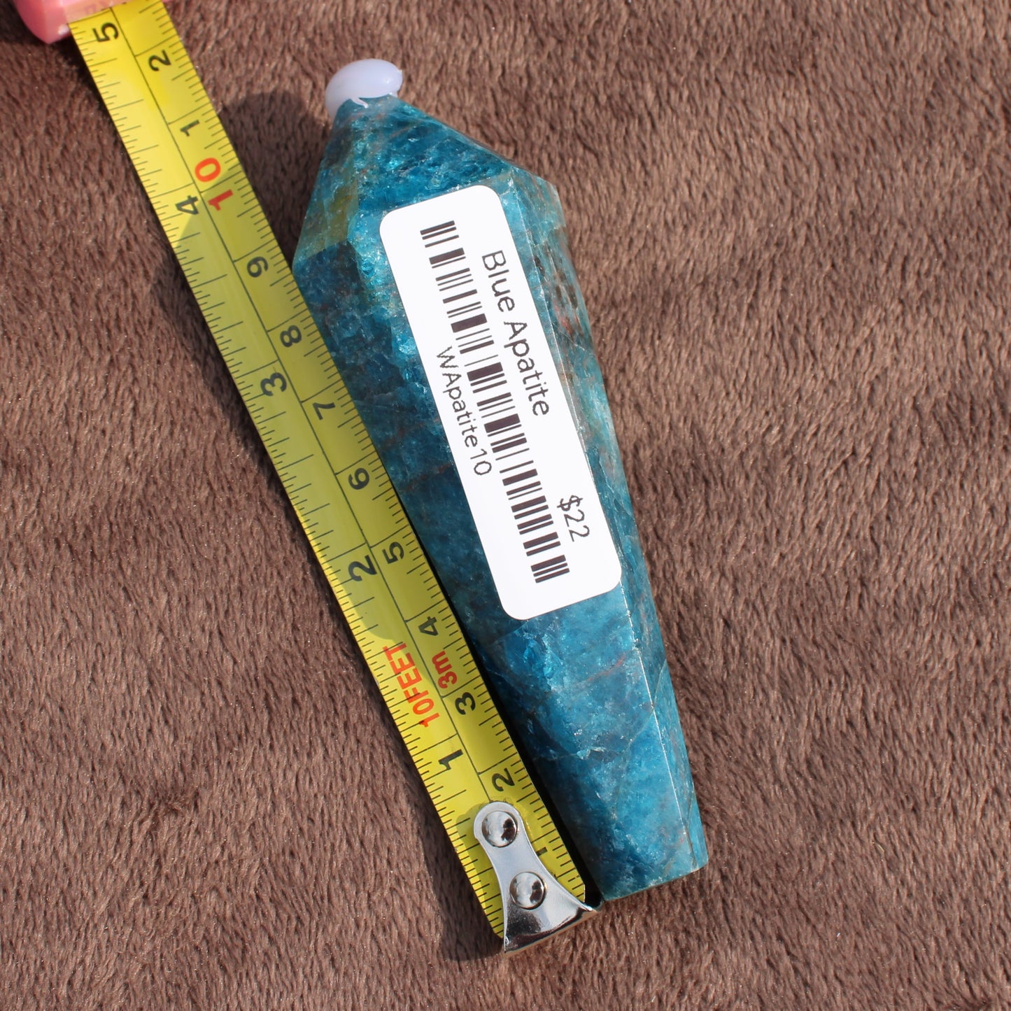 Blue Apatite Wand crystal with price tag next to yellow measuring tape on brown surface