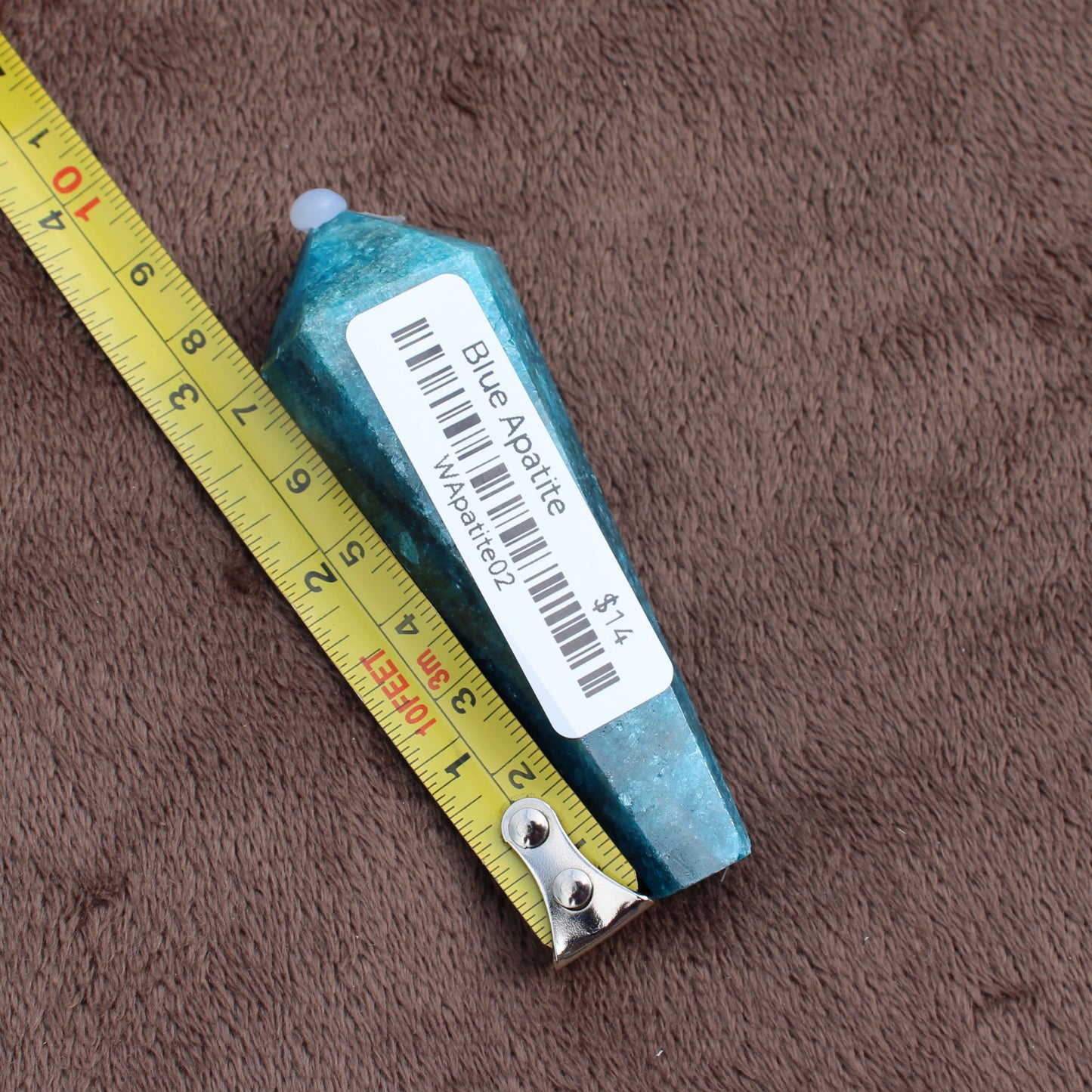 Blue Apatite Wand crystal from Different Branches LLC placed next to a yellow measuring tape on brown fabric