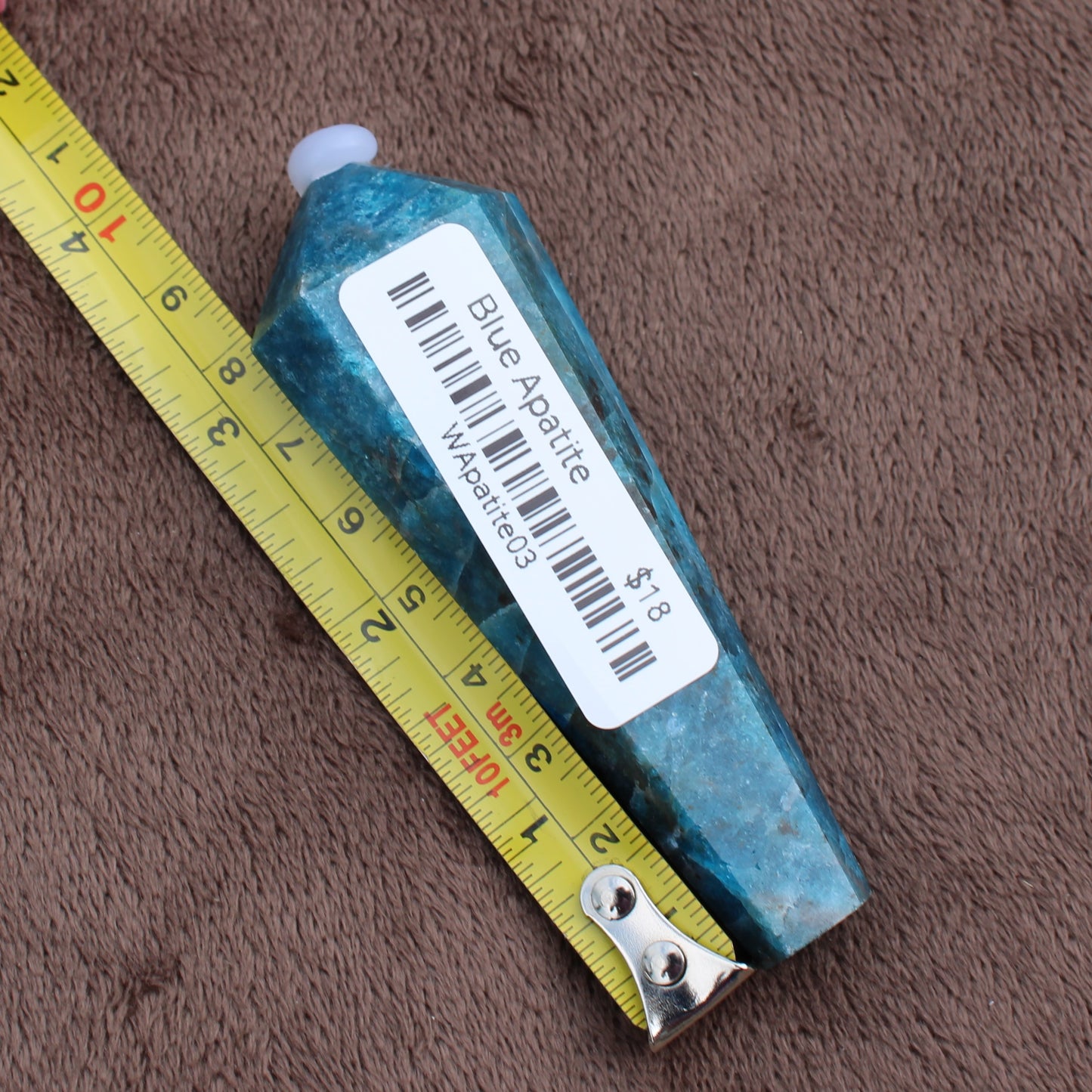 Blue Apatite Wand crystal from Different Branches LLC placed next to measuring tape on brown fabric