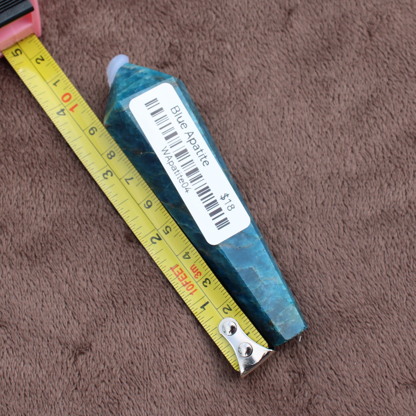 Blue Apatite Wand crystal with price tag next to yellow measuring tape on brown fabric