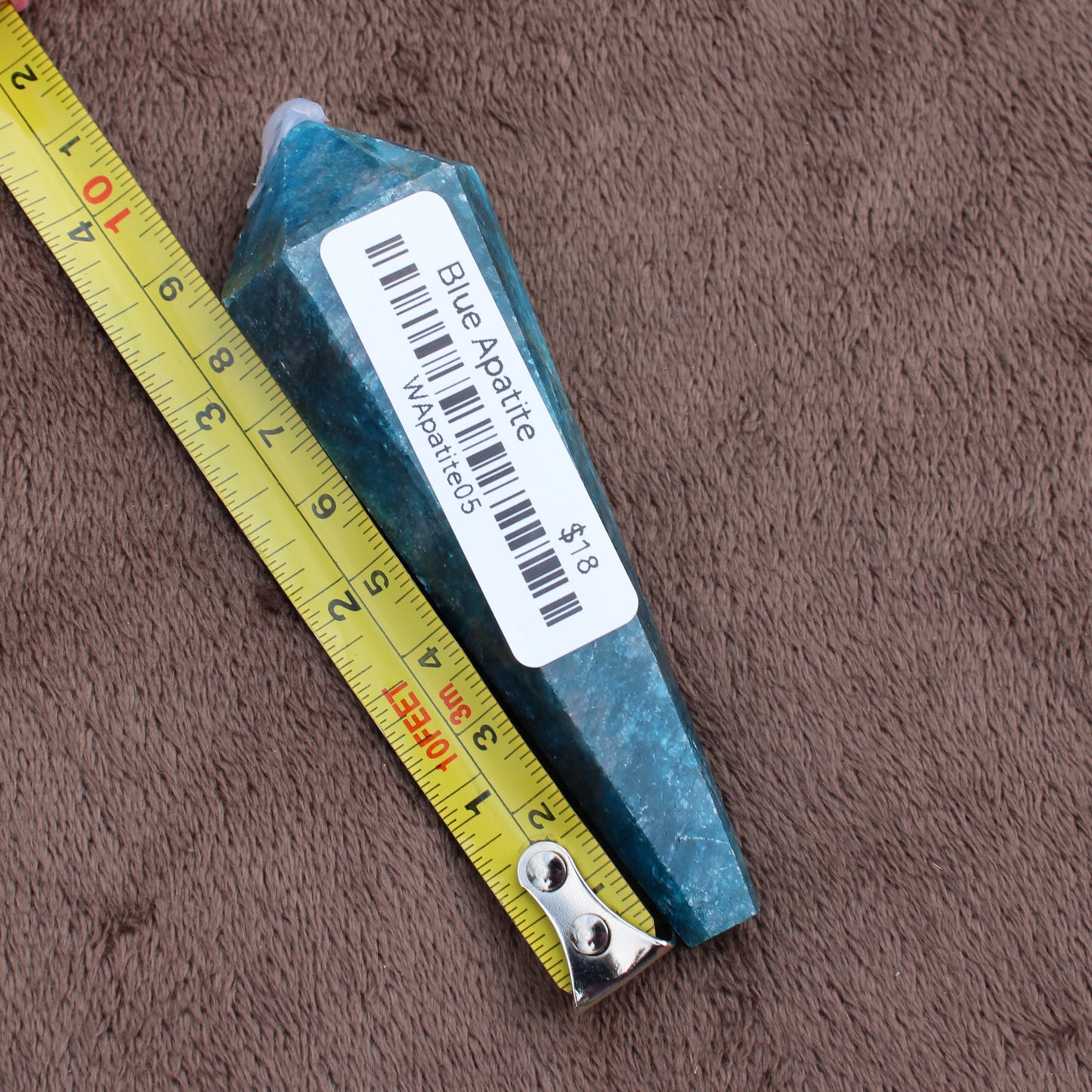 Blue Apatite Wand crystal by Different Branches LLC next to yellow measuring tape on brown fabric