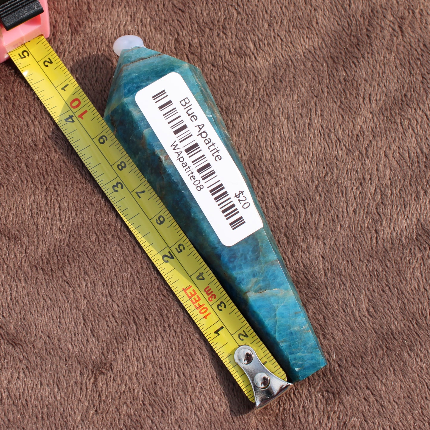 Blue Apatite Wand crystal with label and measuring tape showing size on brown fabric background