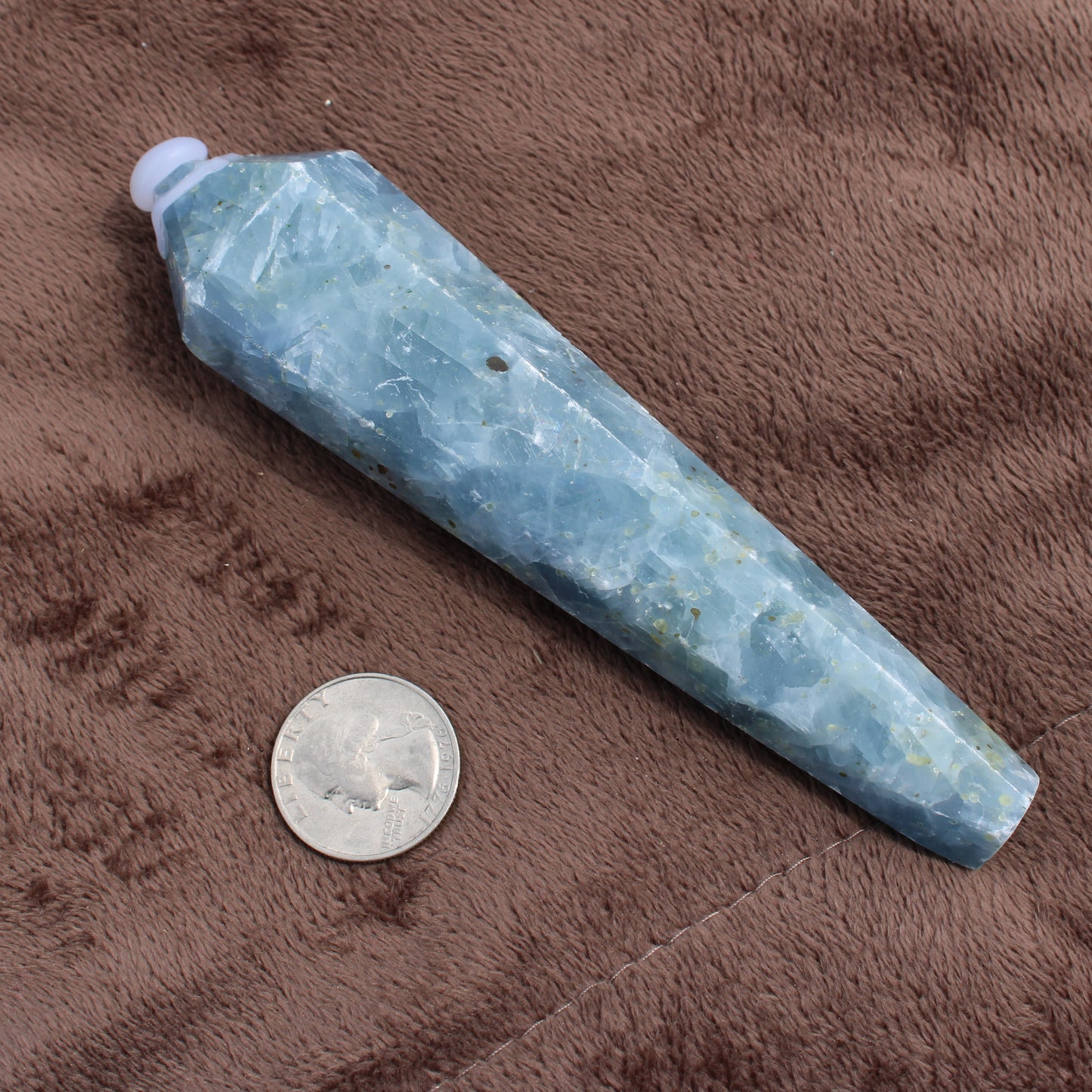 Blue Calcite Wand crystal by Different Branches LLC next to a quarter for size reference on brown fabric