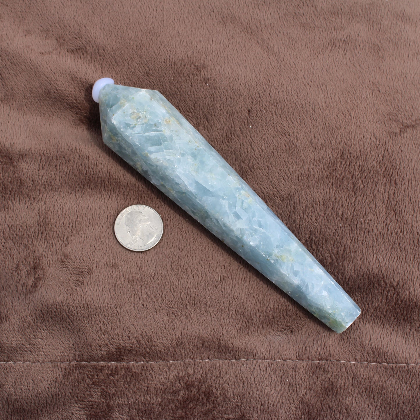 Blue Calcite Wand crystal next to a quarter on a brown fabric background