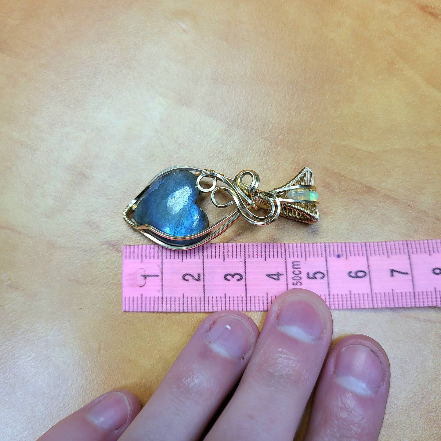 Blue flash labradorite heart pendant set in gold with intricate details and a measuring tape for size reference