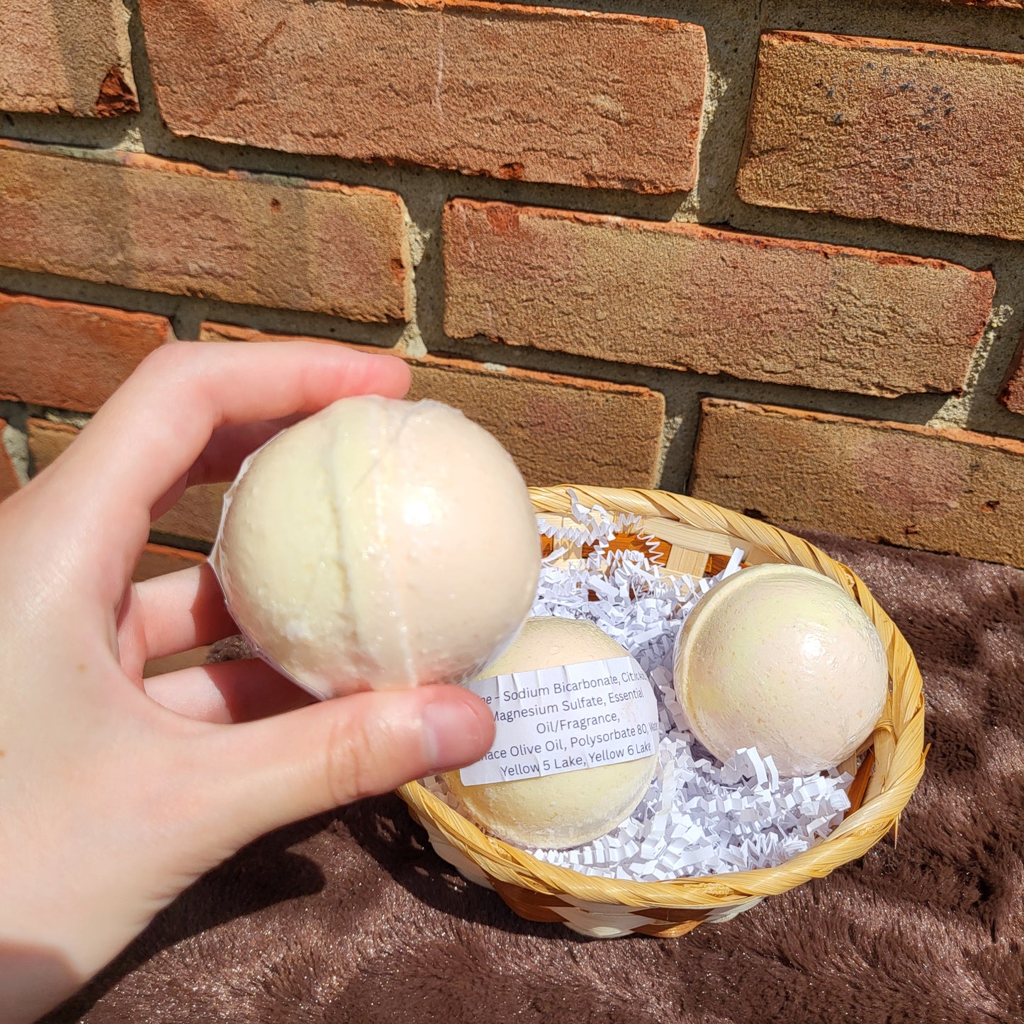Hand holding a Citrine Bath Bomb among a basket of natural bath products against a brick background