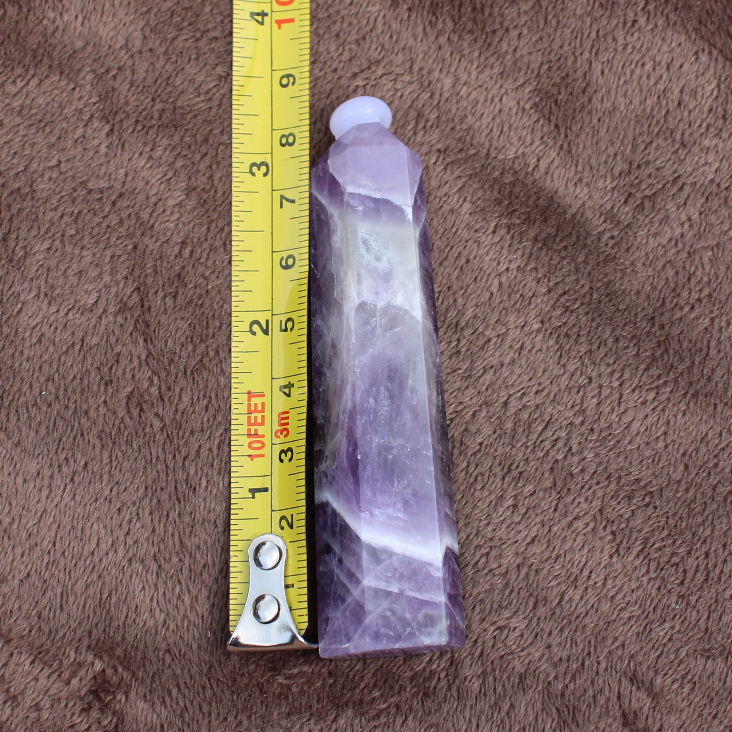 Dream Amethyst Tower crystal measuring about 4 inches placed next to a yellow measuring tape on brown fabric