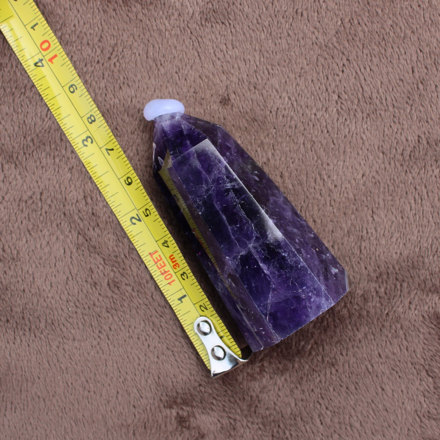 Dream Amethyst Tower crystal measuring about 3 inches next to a yellow tape measure on brown fabric
