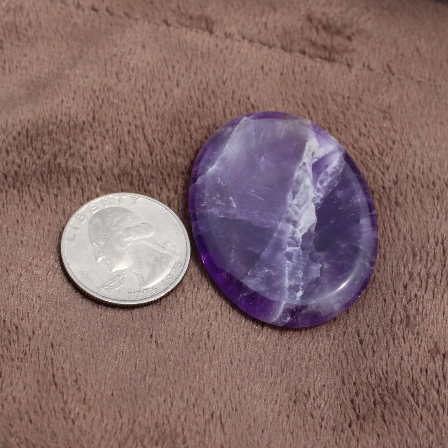 Dream Amethyst Worry Stone from Different Branches LLC next to a quarter for size comparison on a brown fabric surface