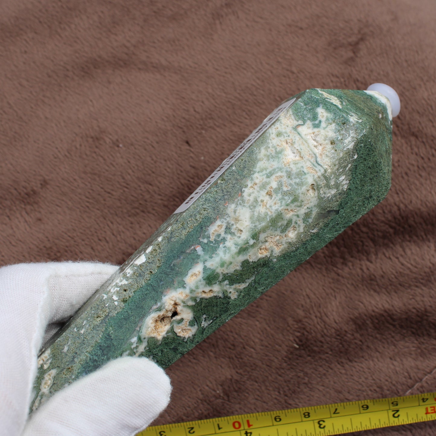 Close-up of a hand holding a polished green and white Moss Agate Wand crystal against a brown background