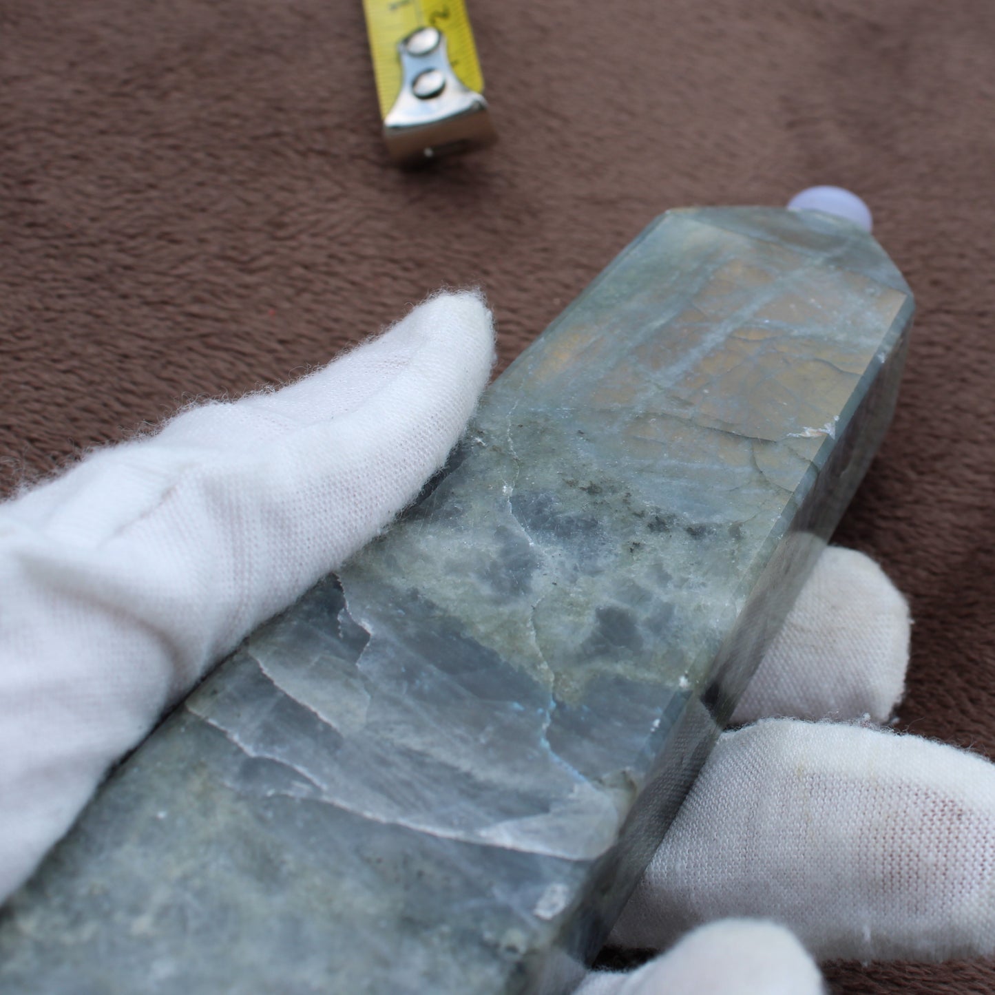 Close-up of a polished Labradorite Tower crystal held by gloved hands showing natural blue and gray hues