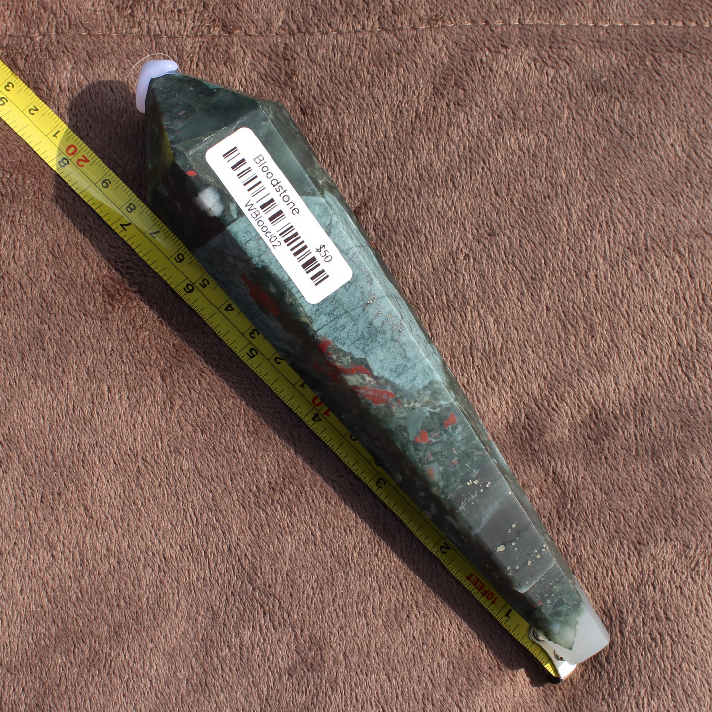 Large Bloodstone Wand crystal with green and red hues displayed with measuring tape on brown fabric