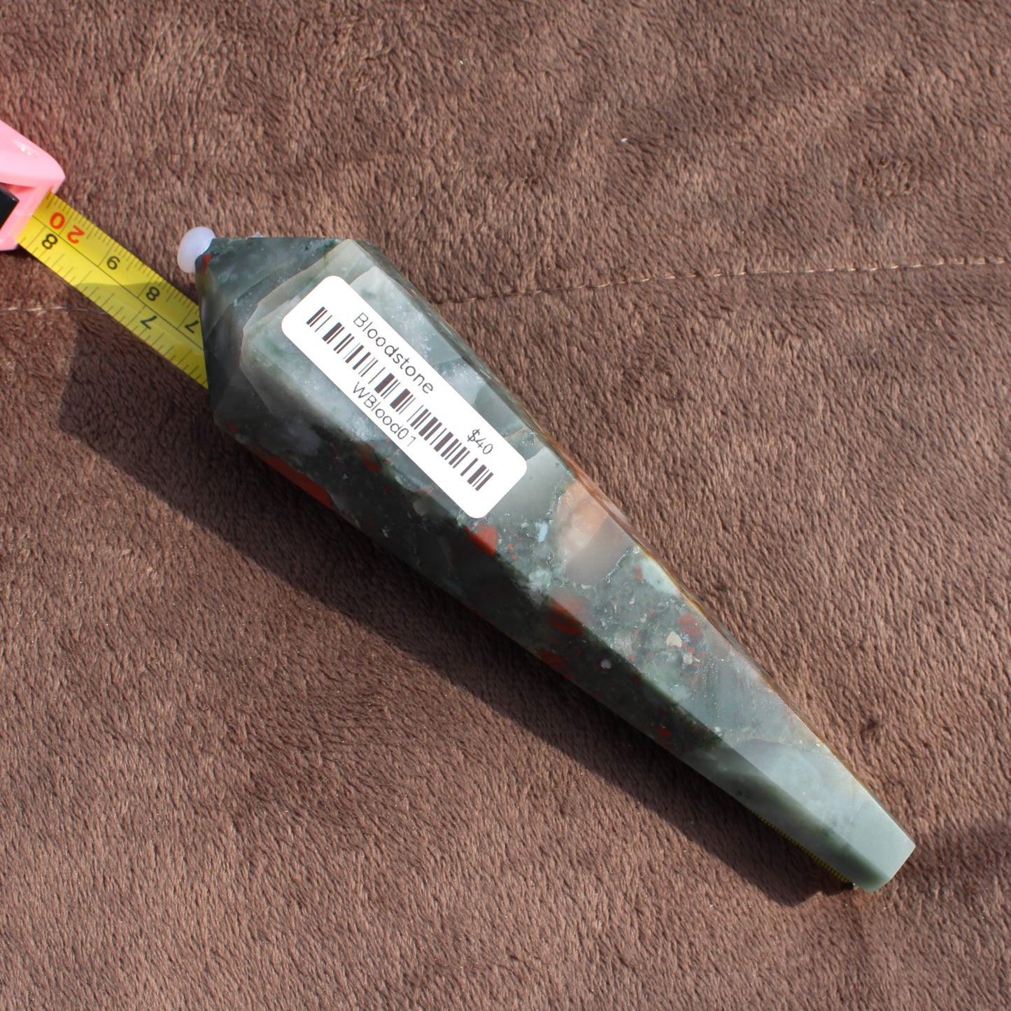 Large Bloodstone Wand crystal with barcode sticker placed next to measuring tape on brown fabric