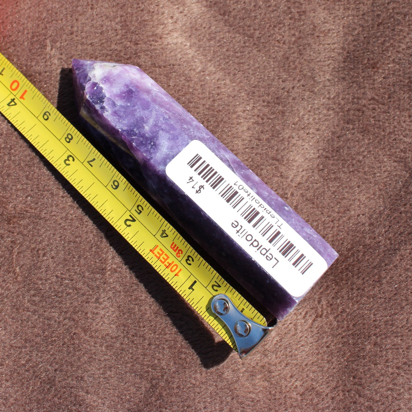 Lepidolite Tower crystal from Different Branches LLC with purple hues next to measuring tape on brown fabric
