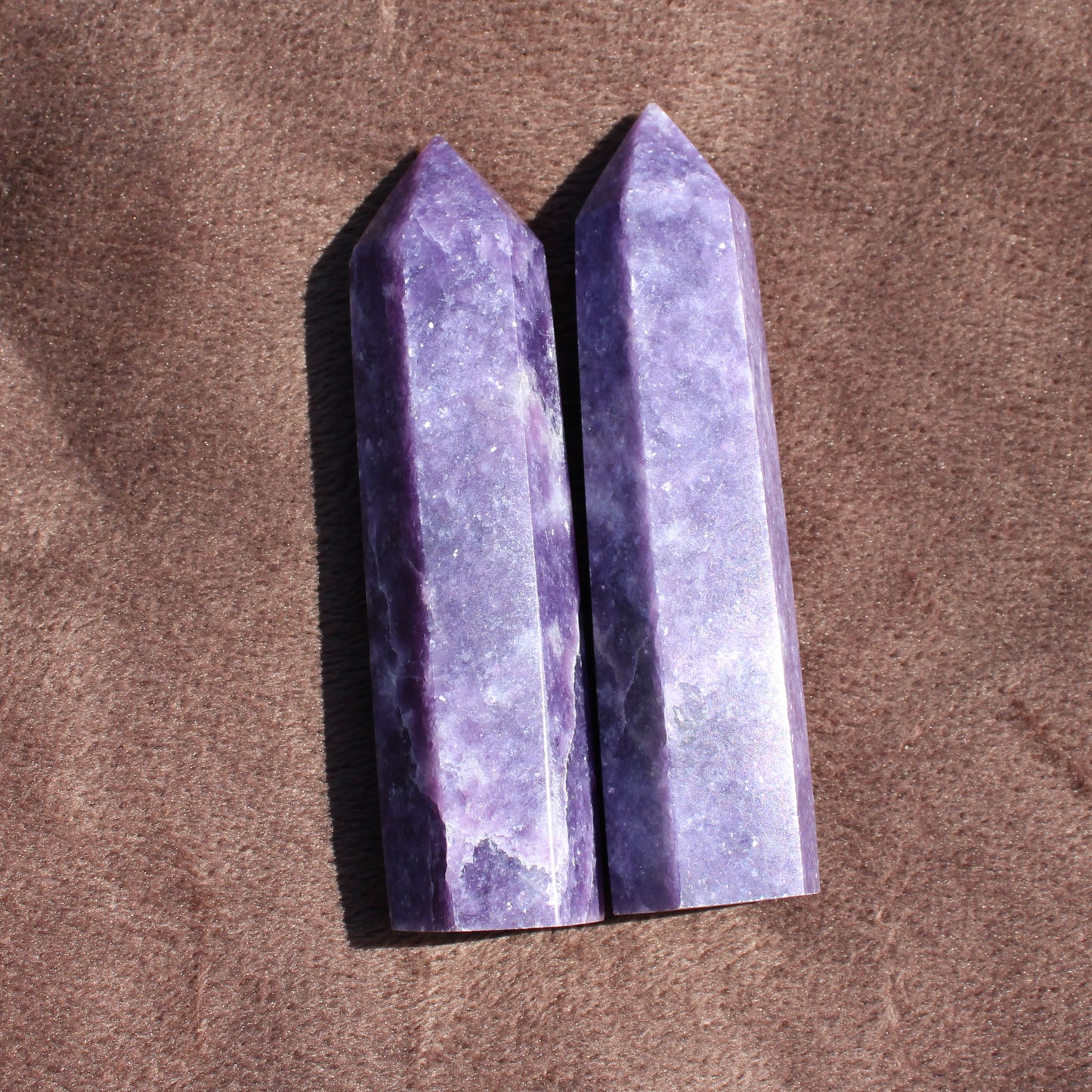 Two polished purple Lepidolite Tower crystals standing upright on a brown fabric background