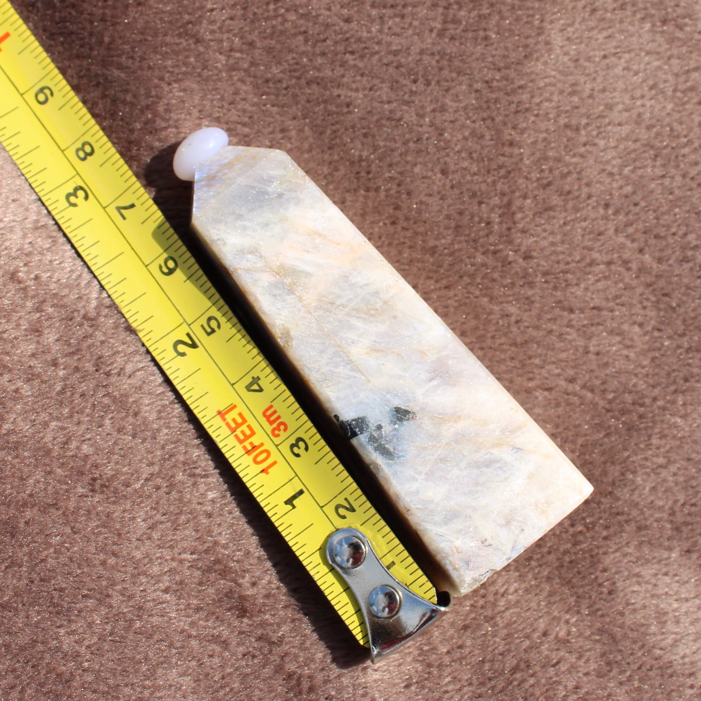 Moonstone Tower crystal from Different Branches LLC next to a yellow measuring tape on a brown surface