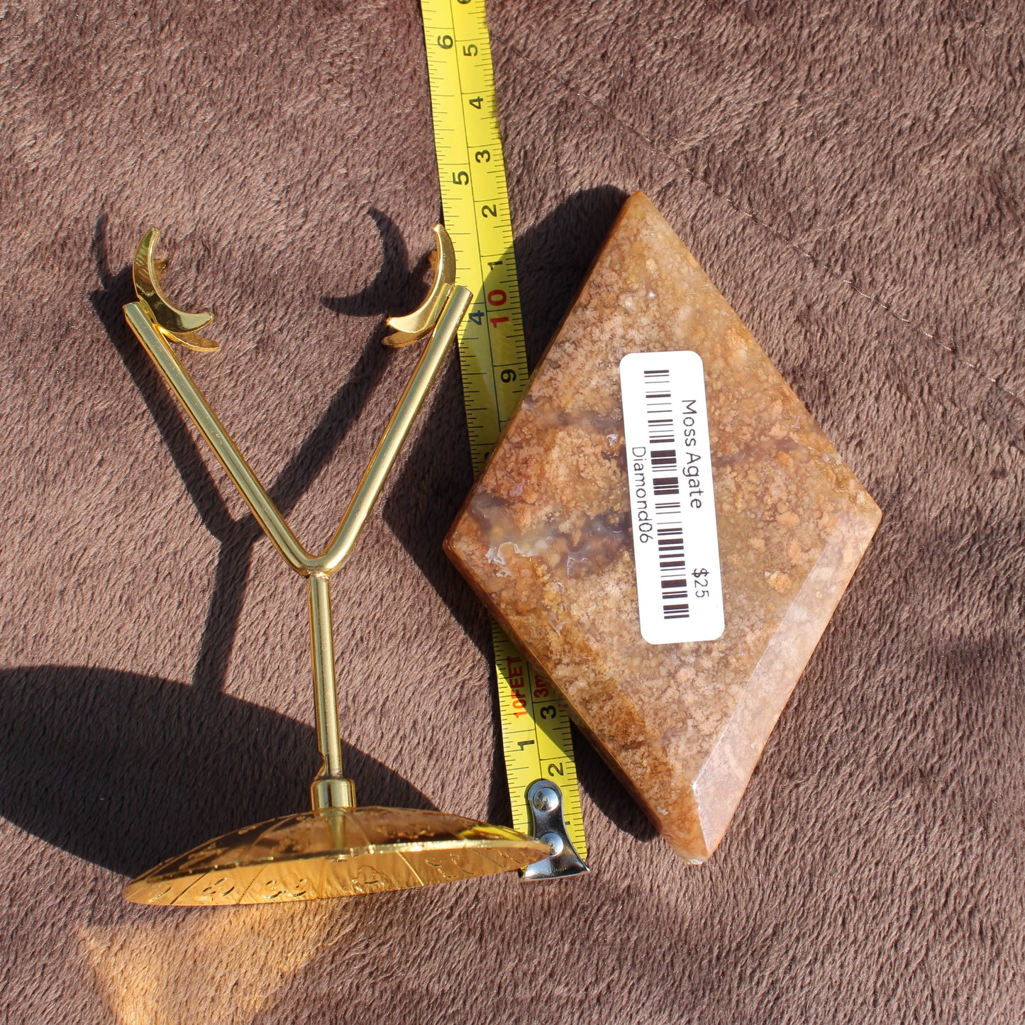 Moss Agate Diamond crystal stone with label next to gold metal stand and measuring tape on brown fabric