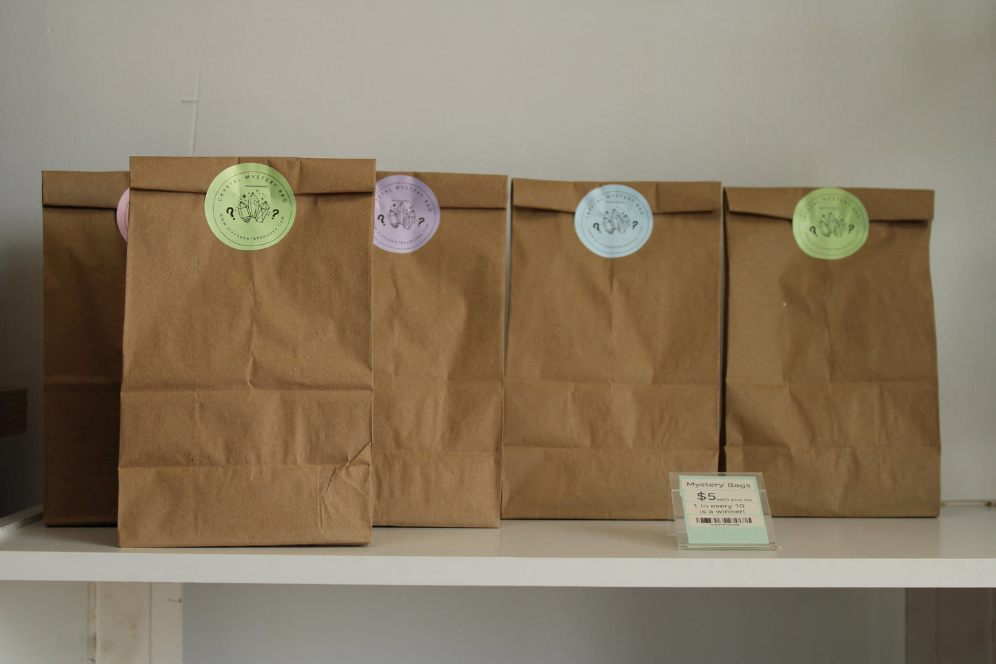 Brown paper mystery crystal bags with colorful round labels displayed on a white shelf