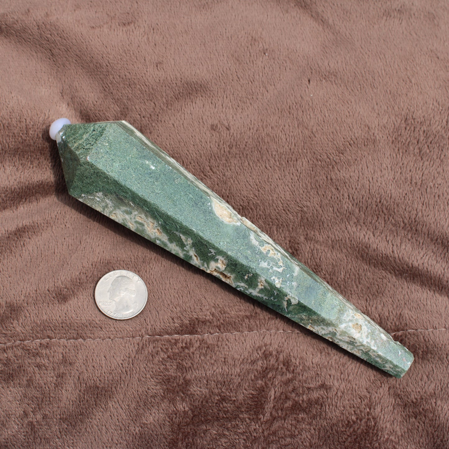 Green and white polished Moss Agate Wand crystal next to a quarter for size comparison