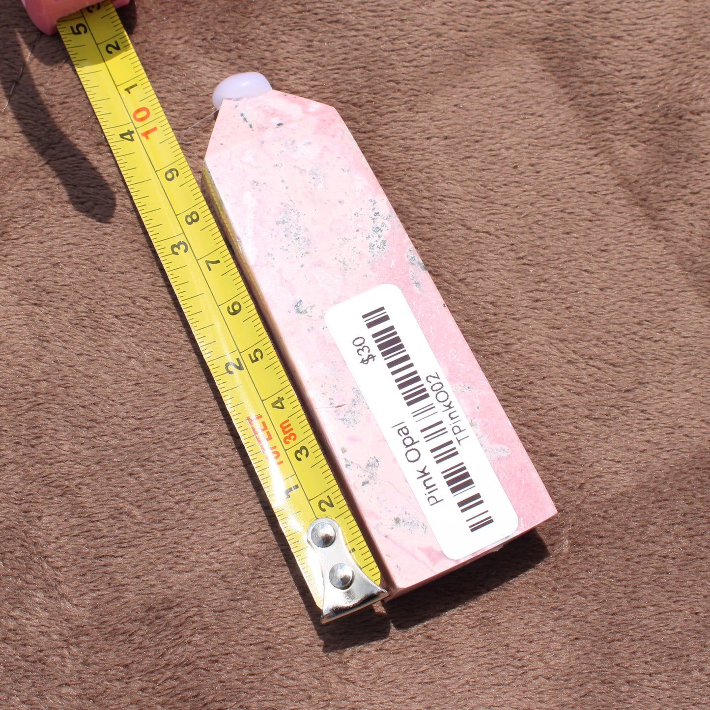 Pink Opal Tower crystal from Different Branches LLC placed next to a measuring tape on a brown fabric background