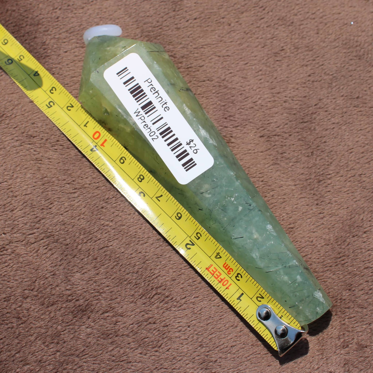 Green Prehnite Wand crystal with barcode label next to yellow measuring tape on brown surface