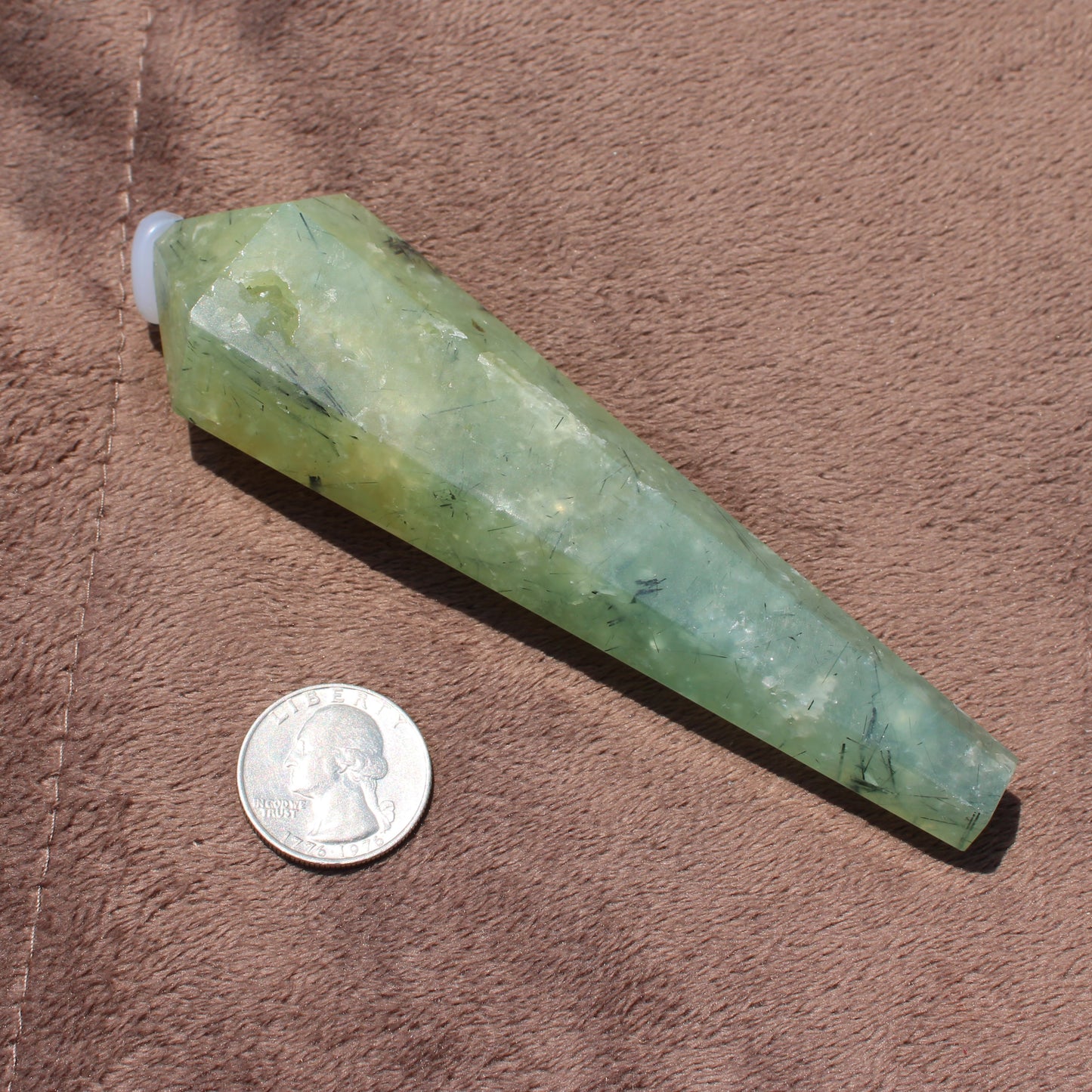 Green polished Prehnite Wand crystal next to a quarter for size comparison on brown fabric