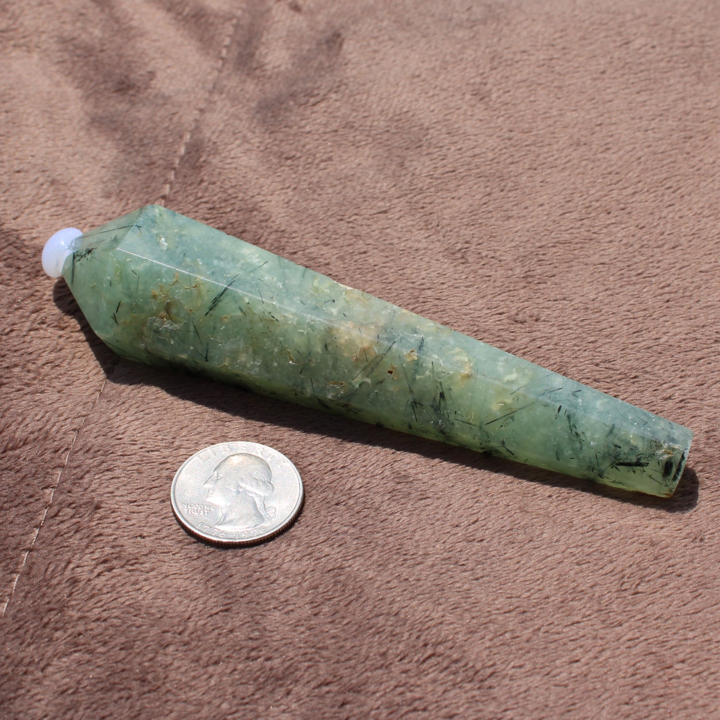 Green Prehnite Wand crystal next to a quarter for size comparison on brown surface