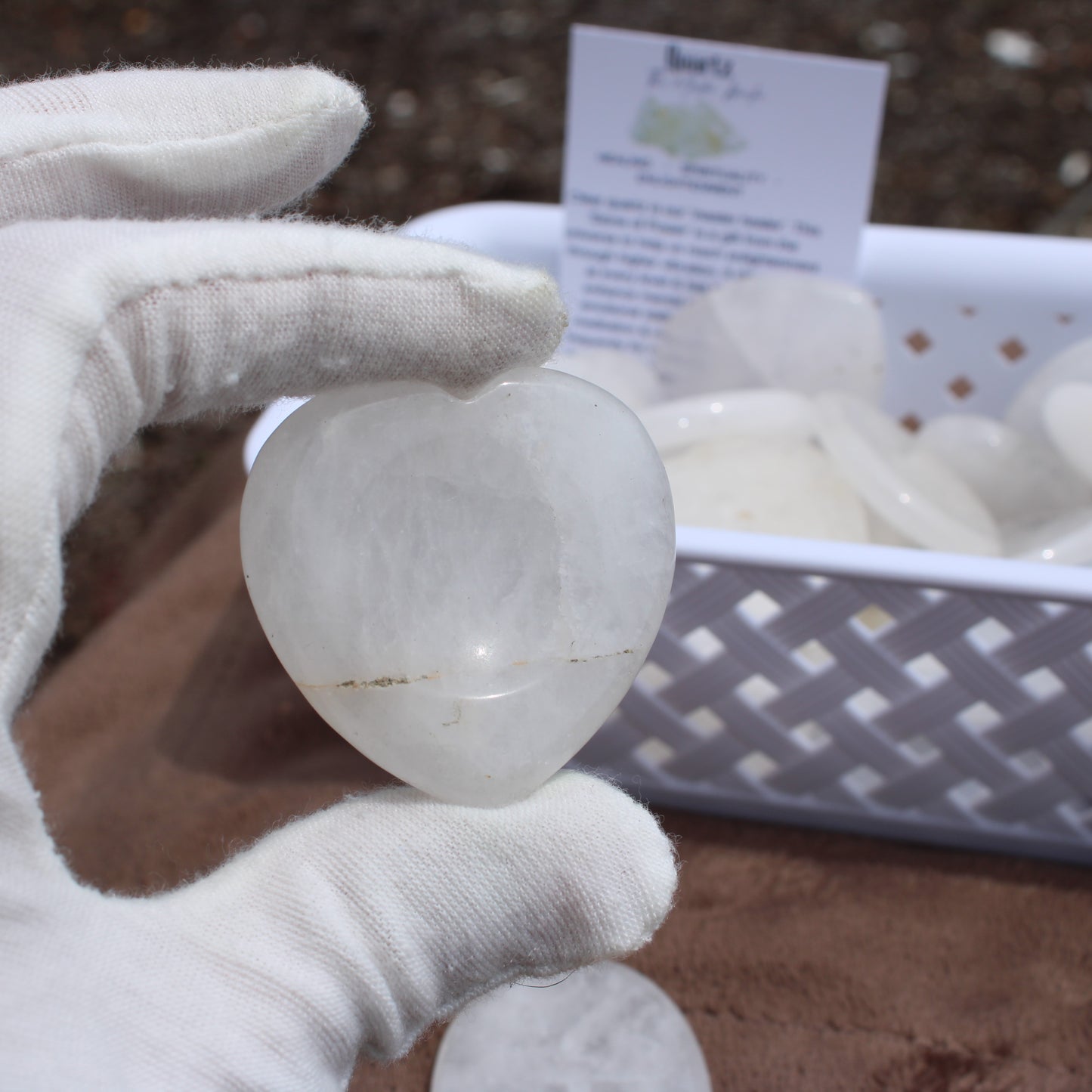 Hand holding a heart-shaped Quartz Worry Stone with white gloves in front of a basket of similar stones