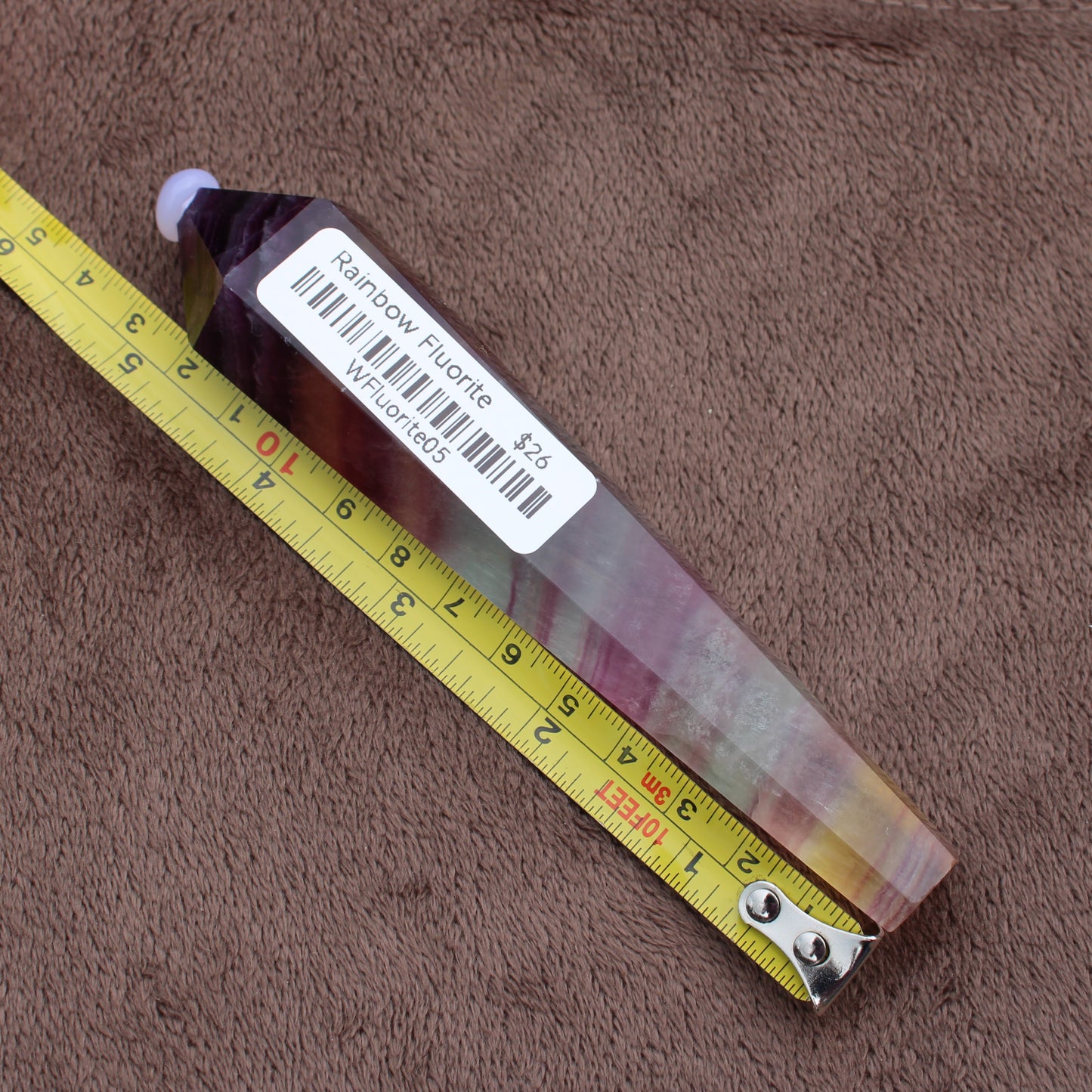 Rainbow Fluorite Wand crystal lying next to a yellow measuring tape on a brown fabric background