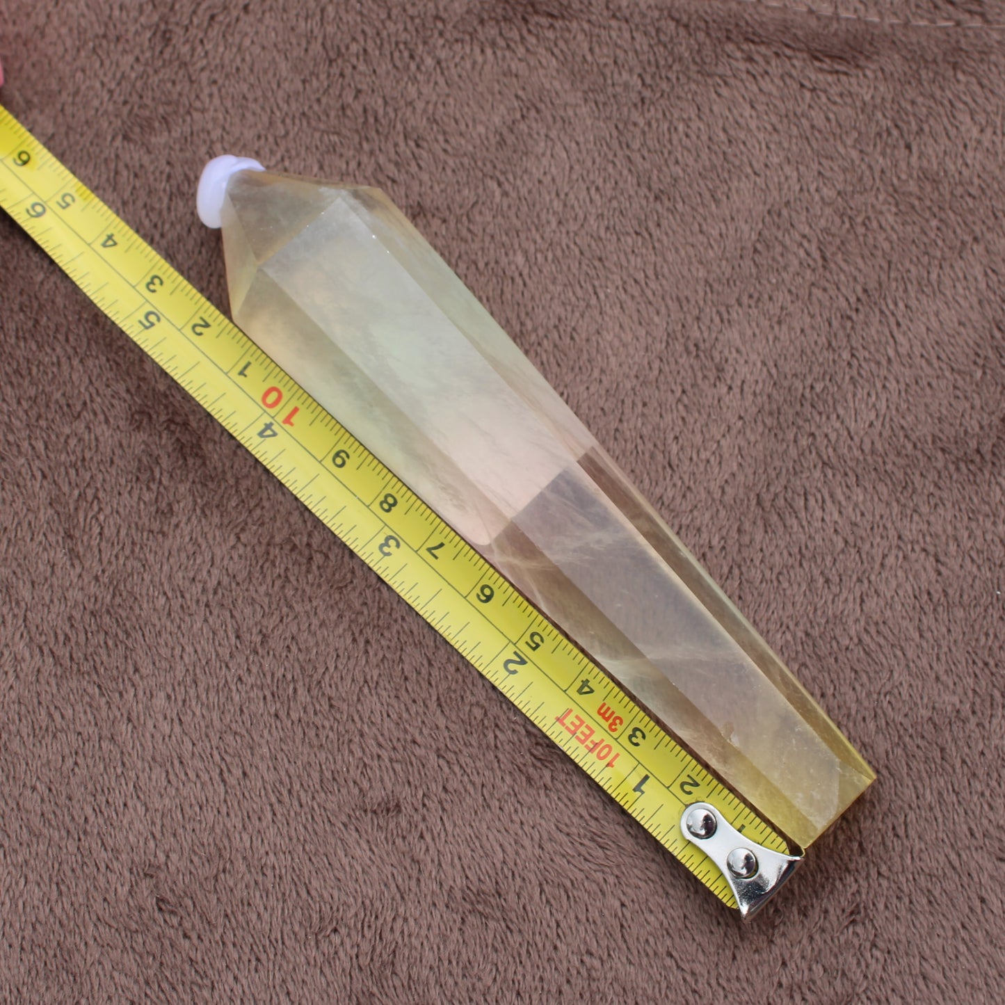 Rainbow Fluorite Wand crystal measuring approximately 8 inches on a yellow tape measure on a brown fabric background