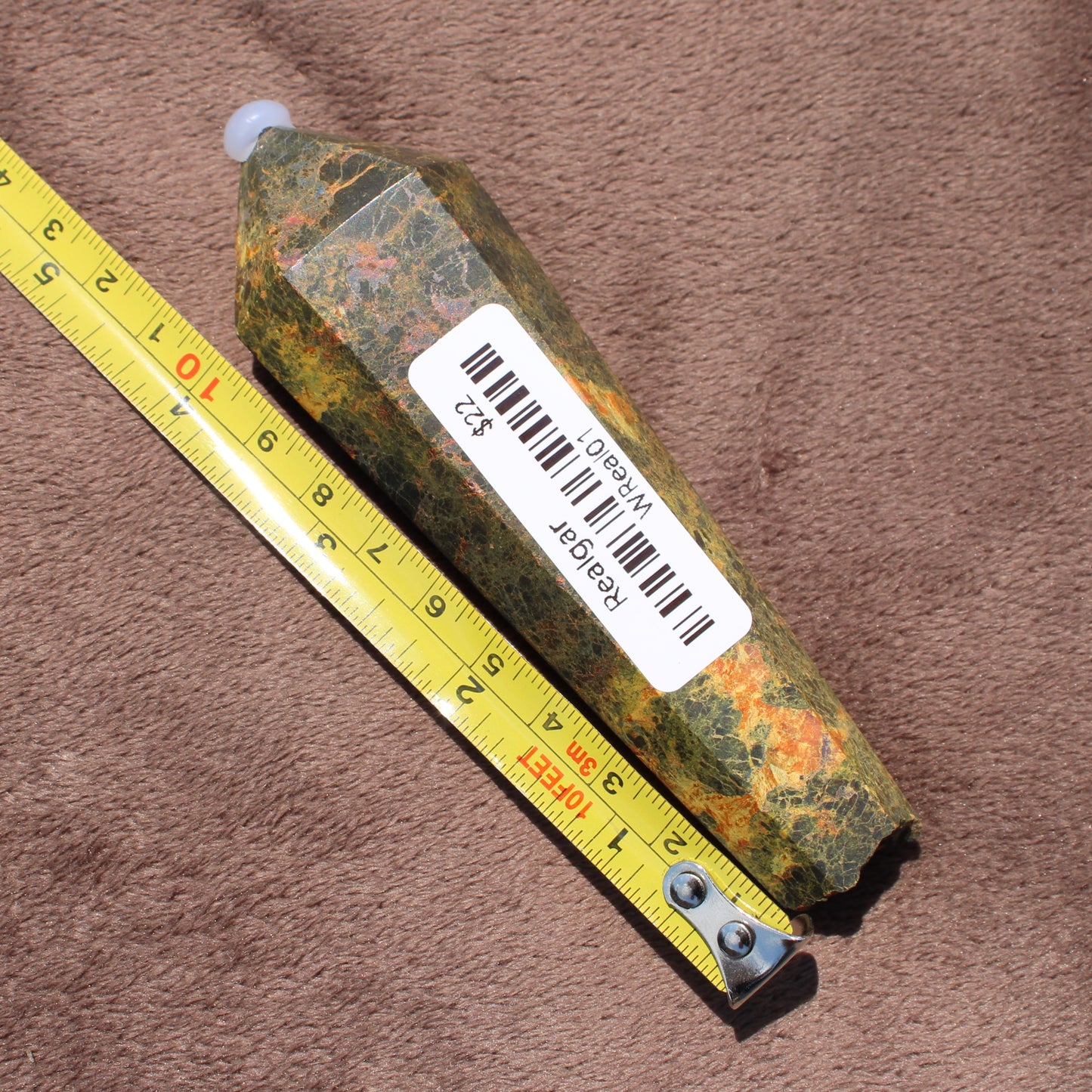 Realgar Wand crystal with green and orange hues placed next to measuring tape on brown fabric