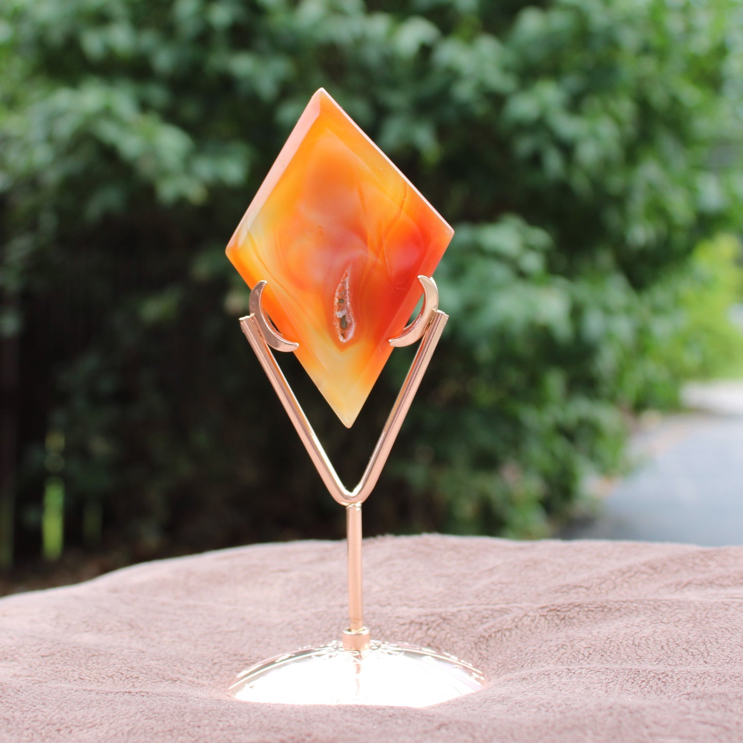 Red Agate Diamond crystal on a rose gold metal stand with a blurred green outdoor background