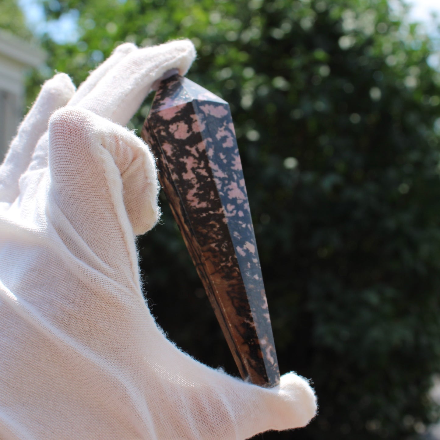 Hand wearing white glove holding a polished Rhodonite Wand crystal outdoors with blurred greenery background