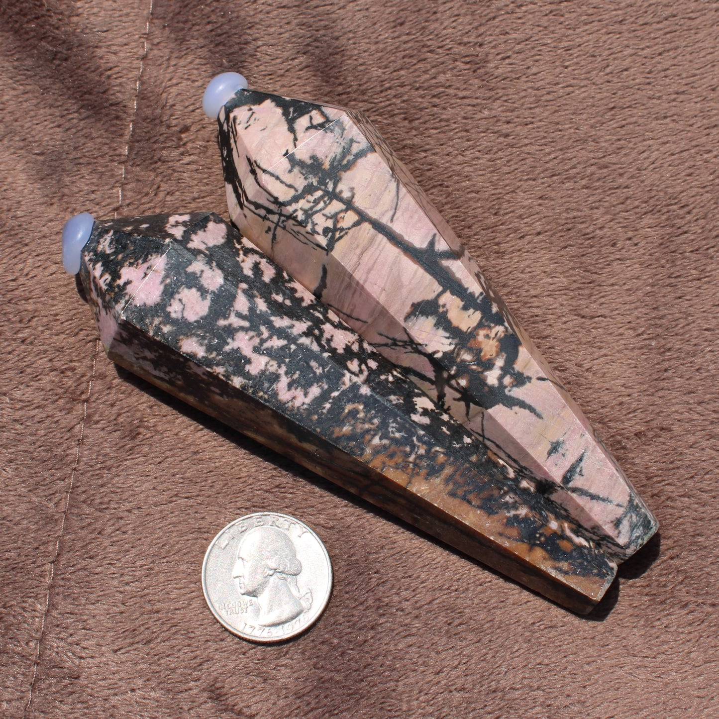 Two polished Rhodonite Wand crystals with pink and black patterns beside a quarter for size comparison