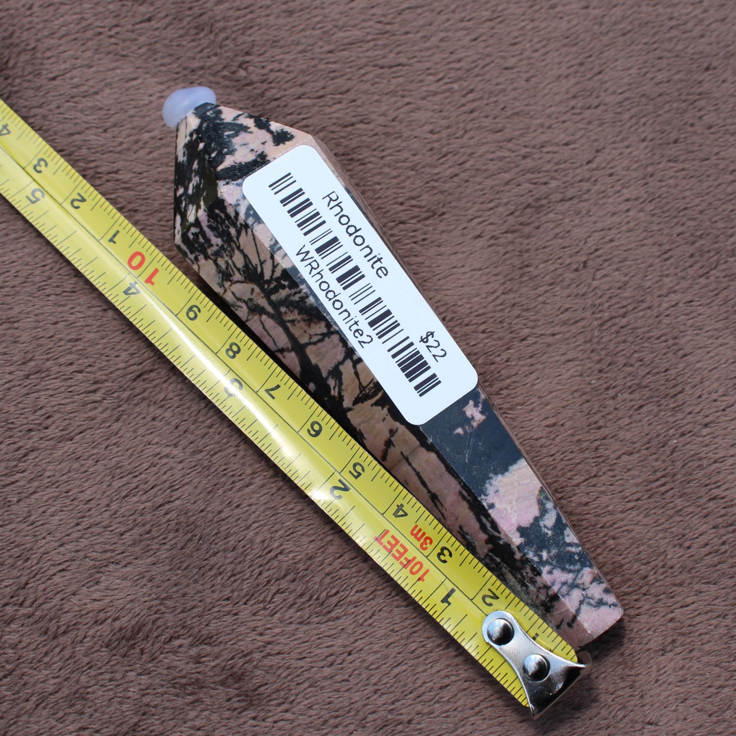 Rhodonite Wand crystal from Different Branches LLC placed next to a yellow measuring tape on a brown surface