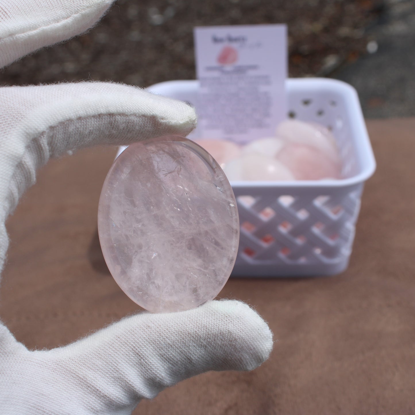 Hand wearing white glove holding a polished Rose Quartz Worry Stone with a basket of similar stones in the background