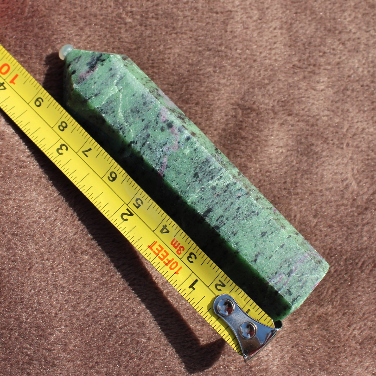 Green and purple Ruby Zoisite Tower crystal point next to a yellow measuring tape on a brown surface