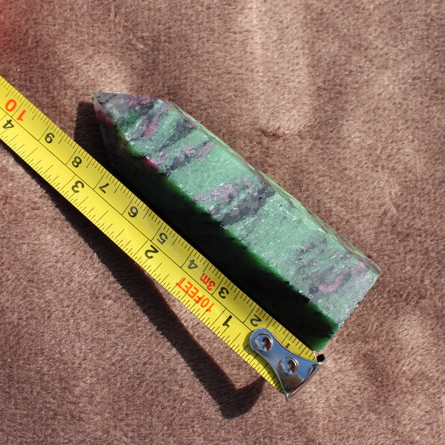 Ruby Zoisite Tower crystal with green and purple hues next to a yellow measuring tape on brown fabric