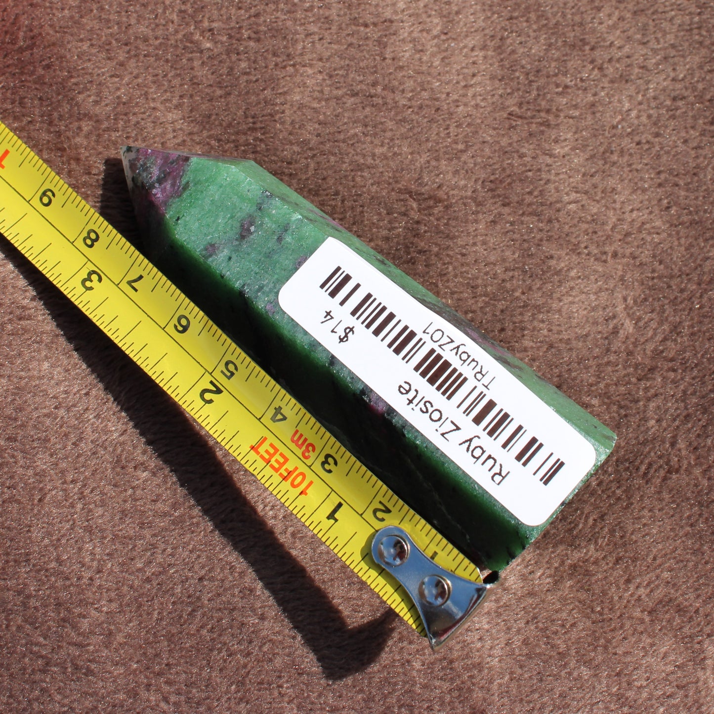 Ruby Zoisite Tower crystal with green and red hues next to yellow measuring tape on brown fabric
