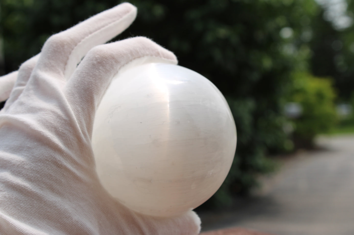 Satin spar selenite sphere held in a gloved hand against a blurred green background