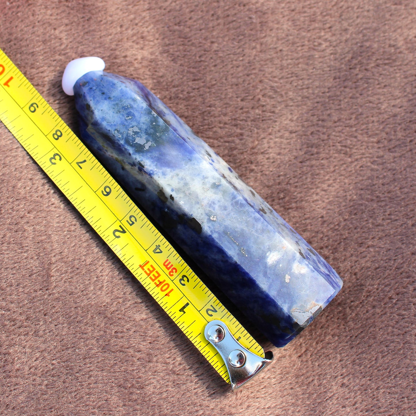 Blue and white polished Sodalite Tower crystal next to a yellow measuring tape on brown fabric