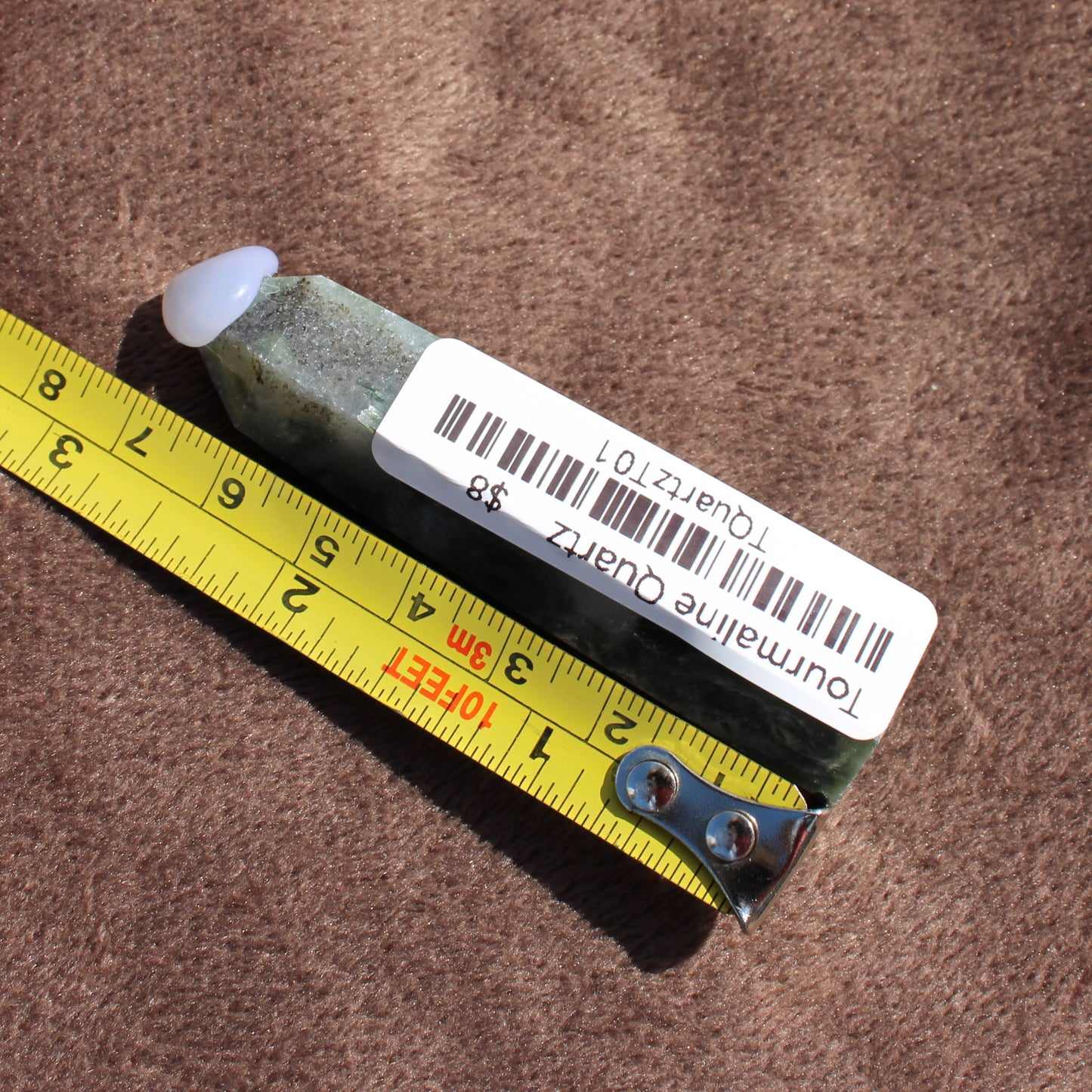 Tourmalited Quartz Tower crystal with barcode label next to yellow measuring tape on brown surface