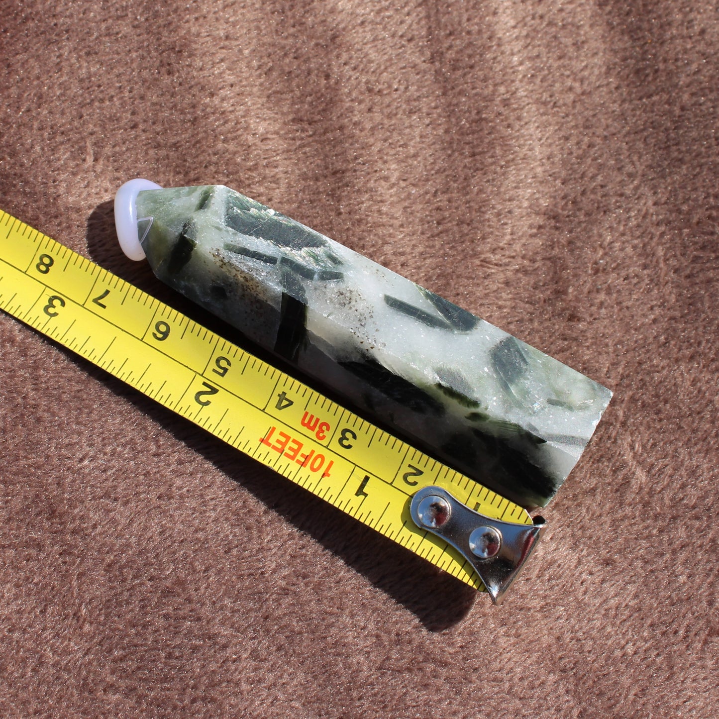 Tourmalited Quartz Tower crystal next to a measuring tape showing its length on a brown fabric background