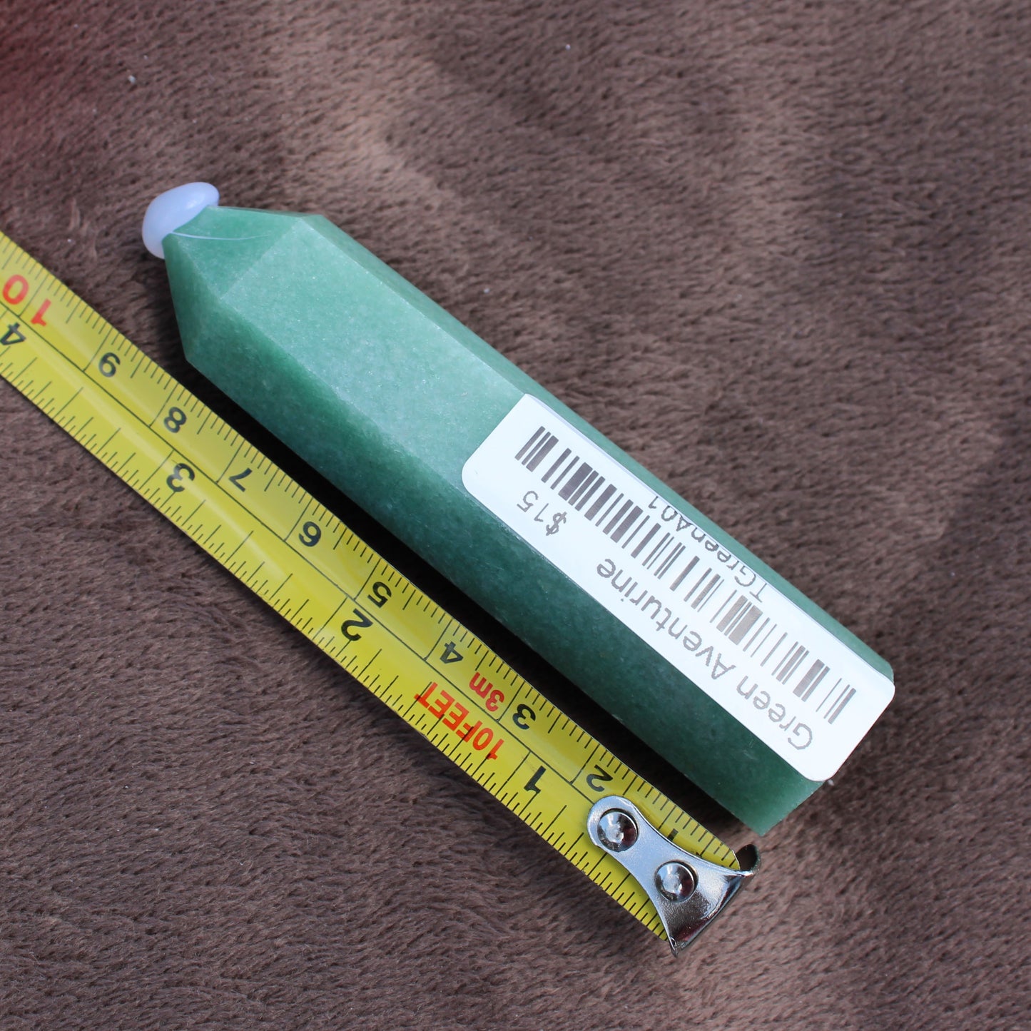 Green Aventurine Tower crystal with barcode label from Different Branches LLC next to a measuring tape on brown fabric