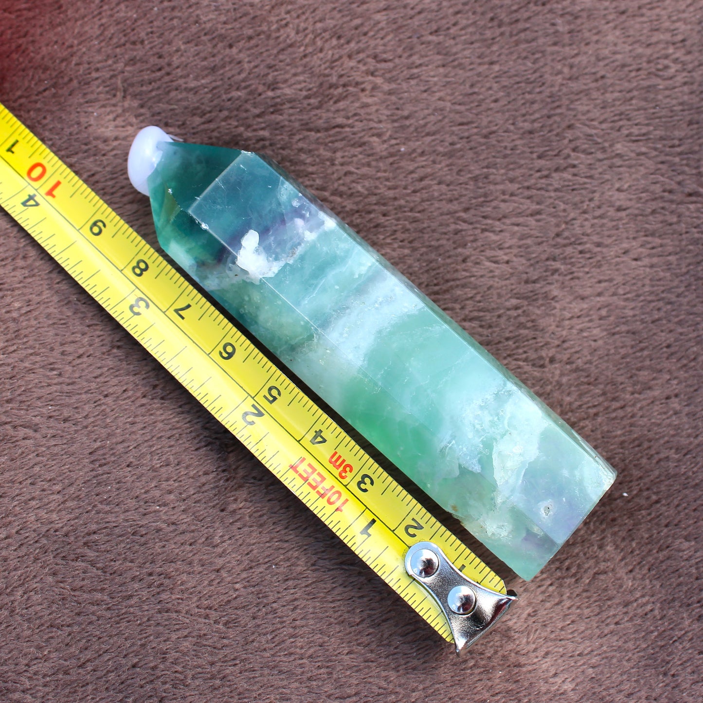 Green Fluorite Tower crystal with polished pointed tip next to yellow measuring tape on brown surface